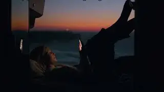 4K Woman lying with legs up and using a phone in an open camper van on the beach at sunset